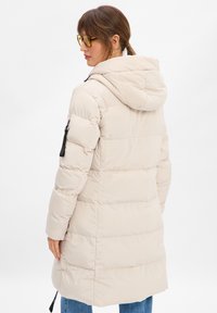 Jasno beżowy puchowy płaszcz z kapturem, z zamkiem błyskawicznym na lewej rękawie i fakturowanym pikowaniem na całej powierzchni.