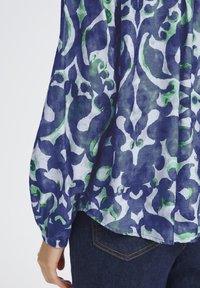 Gros plan sur une personne portant un blouse à motifs floraux bleu et vert avec des manches bouffantes, et une poche de jean bleu foncé visible.