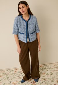 Blauer Tweed-Blazer mit kurzen Ärmeln, rundem Ausschnitt und blauen Akzentnähten; kombiniert mit braunen Hosen mit weitem Bein. Schlichter Hintergrund.