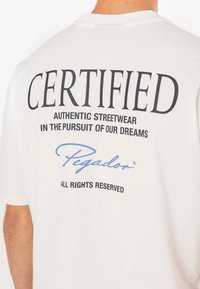 Balta medvilninė marškinė su dideliu juodu tekstu, rašančiu "CERTIFIED", ir mažesniu juodu tekstu žemiau. Yra mėlynas užrašas "Pegador".