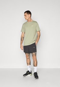 Lichtgroene t-shirt met een klein wit logo, gecombineerd met zwarte shorts met een logo. Zwarte sneakers en witte sokken maken de outfit compleet.