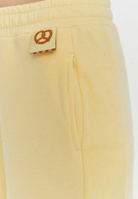 Žluté tepláky mají měkkou tkaninu, elastický pás, boční kapsy a malou nášivku s logem preclíku poblíž pasu.