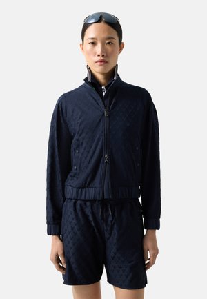 Kobieta w granatowej, teksturowanej kurtce na zamek i dopasowanych szortach, z okularami przeciwsłonecznymi na głowie, stojąca na białym tle.