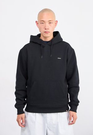 Man with shaved head wearing a black hooded sweatshirt and white pants, standing against a plain white background.