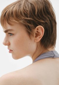 Pendientes de studs en color dorado con un diseño ondulado, llevados por una modelo de cabello corto. El cabello es de color castaño claro, y el fondo es de un blanco neutro.