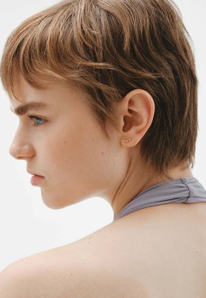 Pendientes de studs en color dorado con un diseño ondulado, llevados por una modelo de cabello corto. El cabello es de color castaño claro, y el fondo es de un blanco neutro.