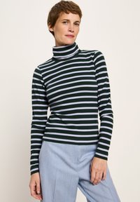 Frau mit kurzen Haaren, die einen schwarz-weiß gestreiften Rollkragenpullover und hellblaue Hose trägt, steht vor einem einfarbigen Hintergrund.