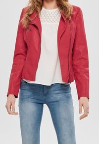 Veste de motard en simili cuir rouge avec fermeture éclair asymétrique, épaules texturées et coupe ajustée, associée à un haut blanc et un jean bleu.