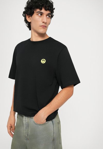 Svart bomulls t-skjorte med rund hals, kortermer, med et lite gult smilansikt-emblem på venstre bryst.