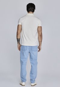 Creme polo-shirt med korte ærmer, parret med lyseblå denim jeans. Skjorten har en afslappet pasform, synlige sømme og kontrasterende krave.