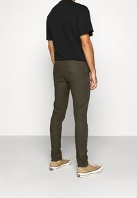 Oliwkowe chinosy o dopasowanym kroju, wykonane z mieszanki bawełny, z detalem tylnej kieszeni, noszone z beżowymi butami i czarną koszulką z krótkim rękawem.
