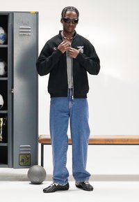 Black zippered bomber jacket over a gray shirt, paired with light blue jeans and black athletic shoes. Accessories include sunglasses and necklace.