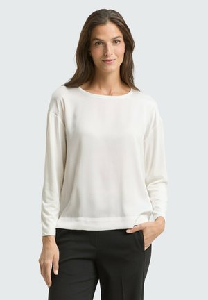 Frau mit langen braunen Haaren trägt eine weiße langärmelige Bluse und schwarze Hose, steht mit einer Hand in der Tasche vor einem schlichten Hintergrund.