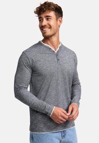 Camiseta henley de manga larga gris con un diseño texturizado, que presenta un collar blanco y una tapeta de botones. Combinada con unos vaqueros de color azul claro.