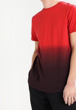 Persoon draagt een T-shirt met korte mouwen met een rood-naar-zwart verloop, met één hand in de zak, staand tegen een effen witte achtergrond.