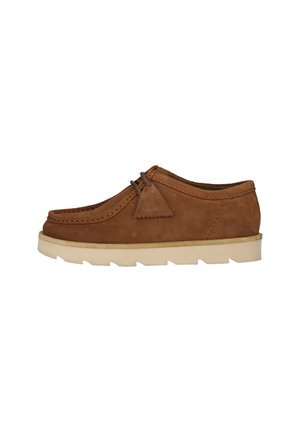 Chaussure décontractée en daim marron avec semelle en caoutchouc blanche et lacets en cuir ornés d'un détail pompon triangulaire sur le côté.