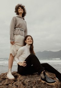 Dos mujeres están de pie sobre una roca junto al océano, vistiendo un suéter beige, una falda crema, jeans negros y botas modernas de tobillo con detalles a rayas.