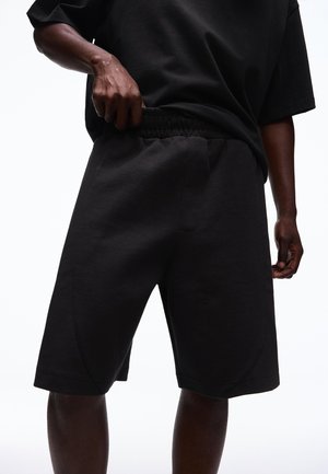 Osoba ubrana w czarne spodenki i czarną koszulkę, poprawiająca elastyczny pas spodenek, pokazana od połowy uda do talii.