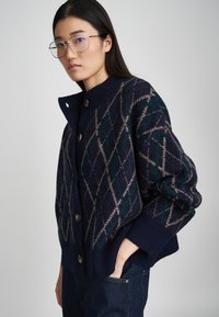 Gilet marin avec un motif en losanges vert et beige, doté de grands boutons marron et d'un col et de poignets en tricot côtelé.
