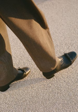 Scarpe in pelle nera con punta arrotondata, tacco piatto e dettaglio a fiocco; abbinate a pantaloni su misura marrone chiaro, presentate su una superficie texture.