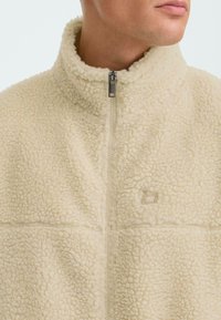 Beige fleecejack met een hoge kraag, halve rits, zachte textuur en subtiele logo-stiksel op de borst. Klassiek, veelzijdig ontwerp.