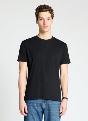 Giovane uomo con capelli ricci che indossa una semplice t-shirt nera a maniche corte e jeans blu, in piedi davanti a uno sfondo chiaro.