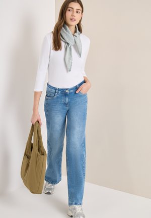Femme portant un haut à manches longues blanc, jean bleu clair, baskets argentées, écharpe grise, tenant un sac cabas vert olive, debout devant un fond neutre.