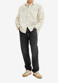 Camicia a maniche lunghe color crema con schizzi grafici, abbinata a jeans neri e sneakers beige. Presenta bottoni e una vestibilità comoda.