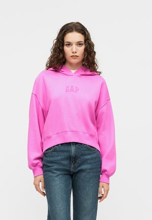 Femeie cu păr creț purtând un hanorac GAP roz aprins și blugi albaștri, stând pe un fundal simplu, deschis la culoare.