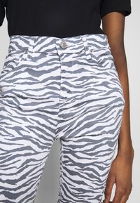 Primer plano de una persona que lleva pantalones blancos con rayas negras de cebra y una parte superior negra, con la mano relajada a su lado.