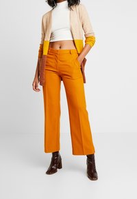 Vrouw met hoogwater mosterdoranje broek, witte cropped coltrui, beige en gele cardigan en donkerbruine laarzen met hak, staand met één hand in de zak.