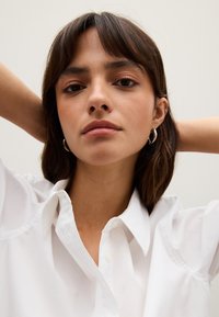 White button-up shirt with a structured collar. Model has long brown hair, wearing silver hoop earrings, with a neutral background.
