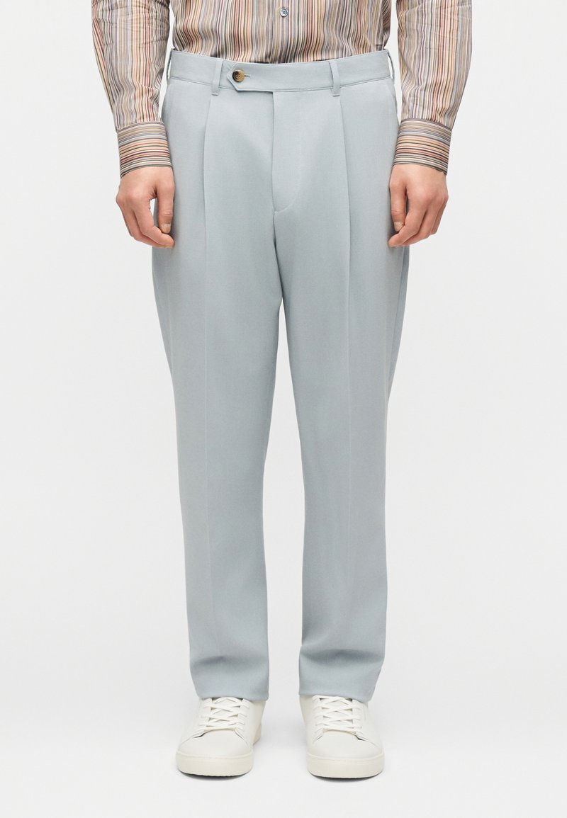 Άνδρας που φοράει ανοιχτό γκρι πλισέ παντελόνι,  λευκά αθλητικά παπούτσια και ριγέ πουκάμισο με μακριά μανίκια, στέκεται μπροστά σε λευκό απλό φόντο.