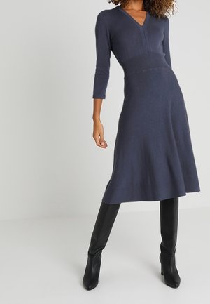 Femme portant une robe midi bleu marine avec un col en V, des manches trois-quarts et des bottes noires montant jusqu’aux genoux avec talons, debout devant un fond uni.