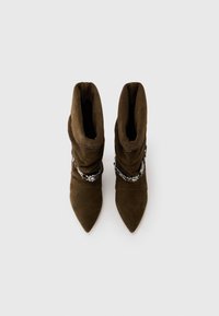 Stivali alla caviglia in suede marrone con design arricciato, punte affusolate e dettaglio in catena argentata sulla parte superiore. Texture liscia con finitura opaca.