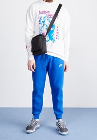 Valkoinen pitkähihainen paita, jossa graafinen kuvio, siniset collegehousut, musta olkalaukku, harmaat tennarit, joissa on teksturoidut yksityiskohdat.