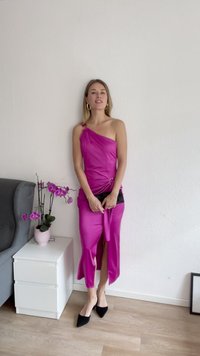 Vienas pleca rozā satīna kleita ar sānu šķēlumu un zelta gredzenu detaļu. Stilizēta ar melniem norādītajiem papēžiem, stāvot priekšā augam un pelēkam dīvānam.