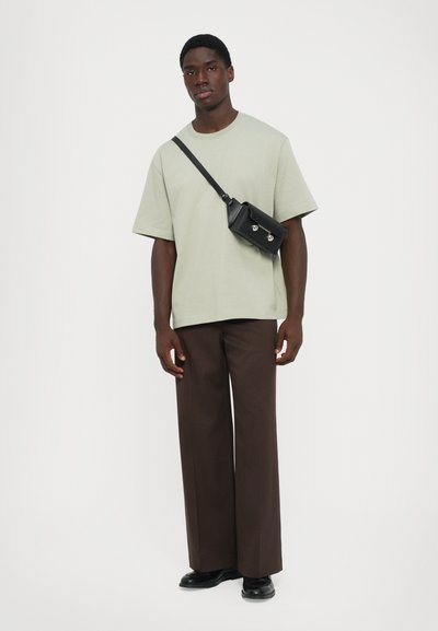 Ung mann iført en lys grønn oversized T-skjorte, brune vide bukser, svarte sko og en svart crossover-veske med sølvdetaljer.