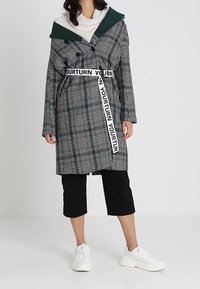 Karierter Trenchcoat in Grün, Schwarz und Weiß mit einer kontrastierenden grünen Kapuze, mit einem Logo-Taillenband gegürtet, kombiniert mit schwarzen cropped Hosen und weißen Sneakers.