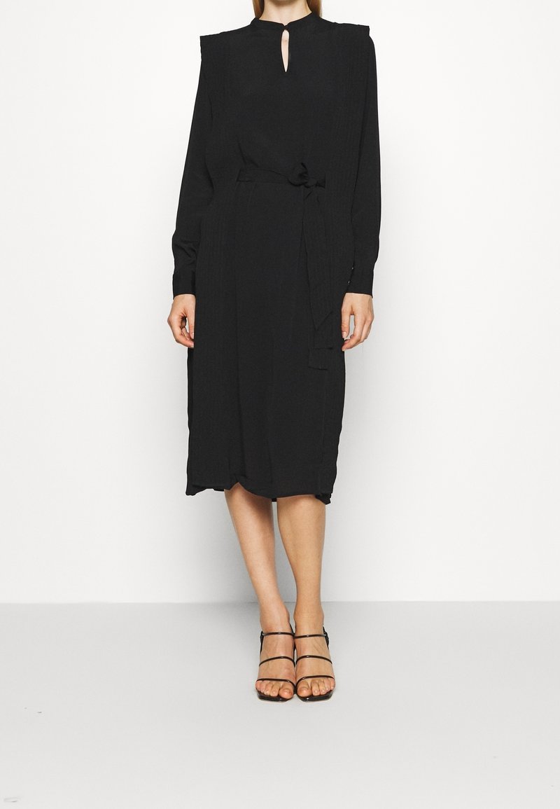 Schwarzes Midi-Kleid mit langen Ärmeln, Schlüssel-Ausschnitt und gebundenem Taillenbereich. Hergestellt aus einem glatten Stoff mit plissierten Schultern.