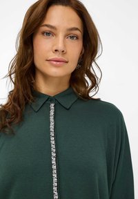 Groene blouse met een zachte textuur, voorzien van een kraag en een centrale knooplijn versierd met heldere kristallen.