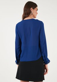Blue long-sleeve blouse with a round neckline, sheer texture, buttoned cuffs, and a curved hem. Paired with a black skirt.