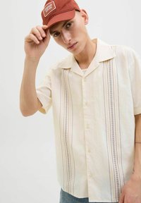 Camisa de manga corta en color crema con rayas verticales multicolores, frente de botones y cuello extendido. Usada con una gorra de color óxido.