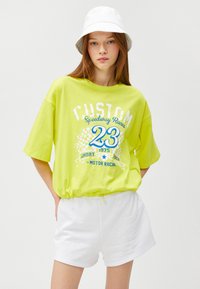 Gul oversized t-shirt med "CUSTOM Speedway Racer" grafik, der har blå accenter og hvid tekst, kombineret med hvide shorts.