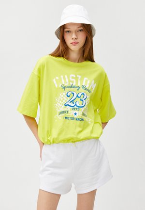 Gul oversized t-shirt med "CUSTOM Speedway Racer" grafik, der har blå accenter og hvid tekst, kombineret med hvide shorts.