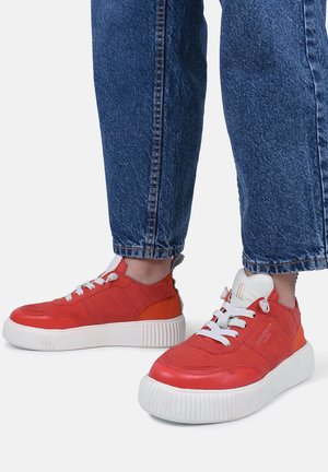 Osoba nosząca czerwono-pomarańczowe tenisówki z białymi grubymi podeszwami i sznurowadłami, w połączeniu z niebieskimi dżinsami.