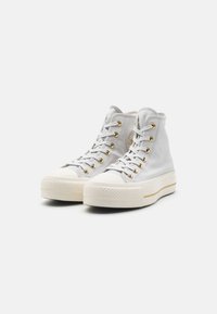 Zapatillas altas de lona gris claro con ojales dorados, puntera de goma blanca y suela texturizada. Presentan un logo en el lateral.