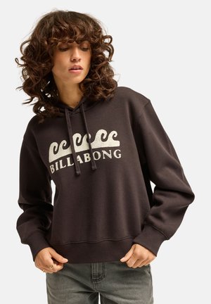 Donna con i capelli ricci che indossa una felpa nera Billabong con il logo a onda, in piedi con le mani vicino alla vita, guarda in basso verso sinistra.