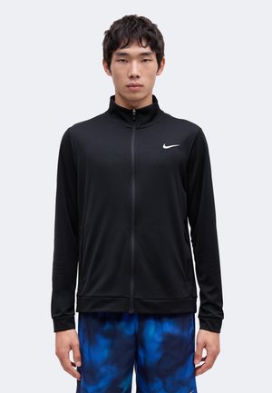Mladý muž v čiernej Nike mikine na zips a modrých vzorovaných šortkách stojí pred jednoduchým svetlým pozadím.