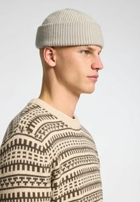 Un berretto a costine di un grigio chiaro indossato da una persona calva, abbinato a un maglione crema e marrone con motivi geometrici.